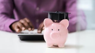 Businessperson's Hand Using Calculator Beside Piggybank With Graduation Hat