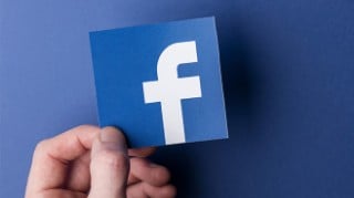 Close up photo of a left hand holding a cut out of the Facebook logo over a blue surface.