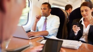 Commuters sitting at a table aboard a train looking at laptops and smartphones.