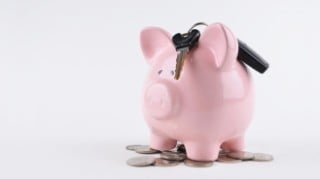 Pink piggy bank with car keys balanced on top of it, standing on a pile of silver coins.