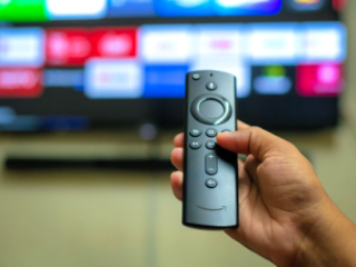 Close up of a hand pointing an Amazon Fire Stick remote control at a television.