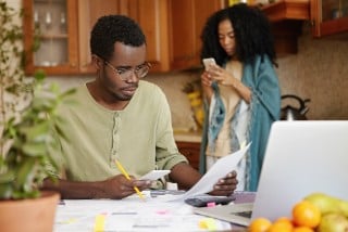 Serious and concentrated young African-American male wearing spectacles paying bills online, using banking app on his mobile phone. People, paperwork, finances, family budget and economy concept