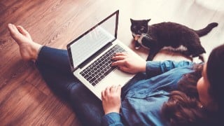 Young woman sitting on wooden floor typing on a laptop on her lap with a black and white cat laying to her right.