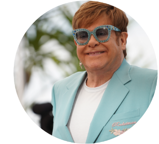 Elton John smiling while wearing light blue glasses and jacket.