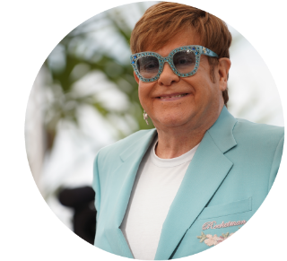 Elton John Elton John smiling while wearing light blue glasses and jacket.