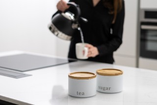 Person pouting water from a stove-top kettle into a mug, with sugar and tea containers in the foreground