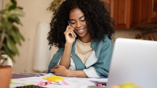 Portrait of beautiful young African housewife with braces smiling happily, talking on phone while sitting at kitchen table with calculator and laptop pc, managing family budget and doing paperwork